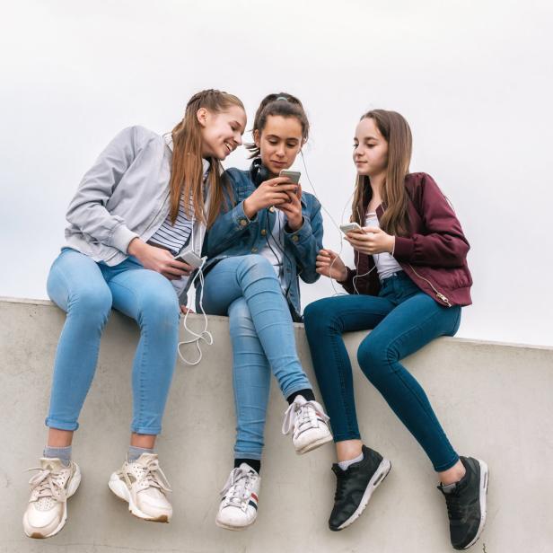 Drei Teenager sitzen auf einer Mauer und schauen auf ihre Handys.