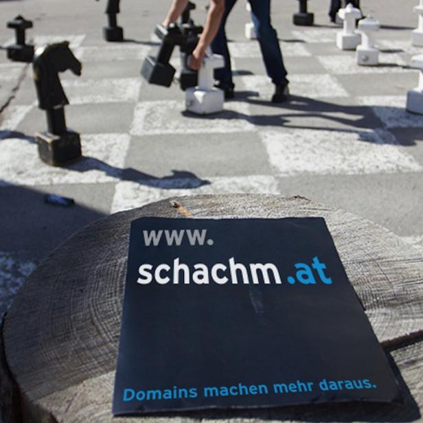 Eine Person bewegt eine Schachfigur auf einem übergroßen Schachbrett im Freien. Im Vordergrund liegt ein Flyer für schachm.at.