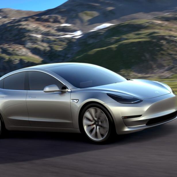 Ein silbernes Tesla Model 3 fährt auf einer Straße vor einer Bergkulisse.
