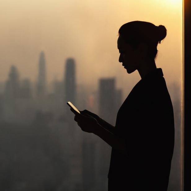 Silhouette of young woman using smartphone next to window with cityscape