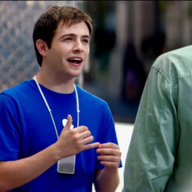Ein junger Mann in einem blauen T-Shirt mit Apple-Logo spricht mit einer anderen Person.