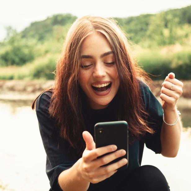 Young smiling woman sitting next to a river typing messages on her smart phone