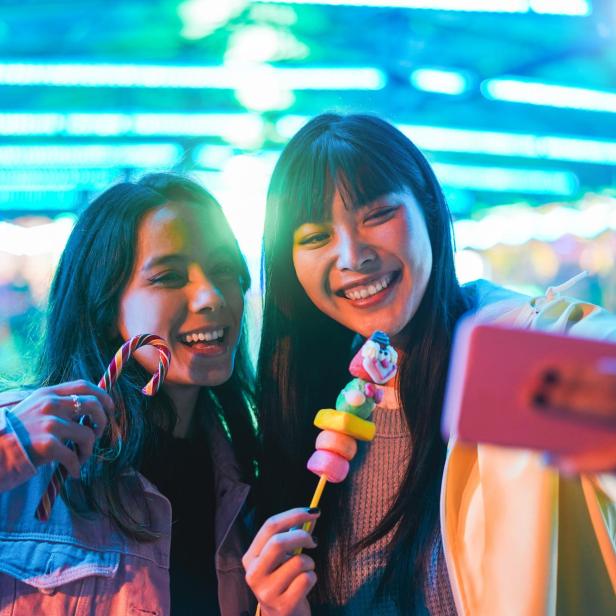 Happy asian girls eating candy sweets and taking selfie at amusement park - Young trendy friends having fun with technology trend - Tech, friendship and influencer concept - Focus on right female face