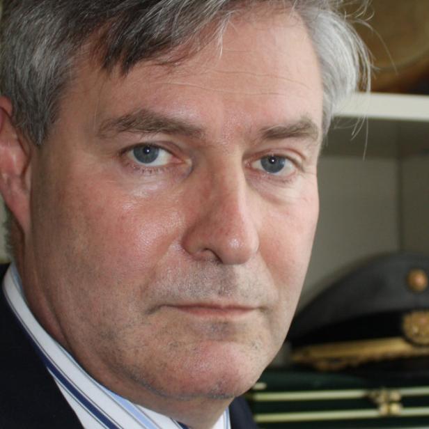 Porträt eines Mannes in Anzug und Hemd vor einem Regal mit einer Militärmütze.