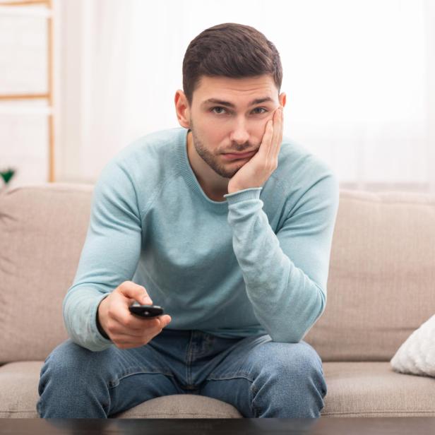 Ein Mann sitzt gelangweilt auf einem Sofa, hält eine Fernbedienung und stützt den Kopf auf die Hand.