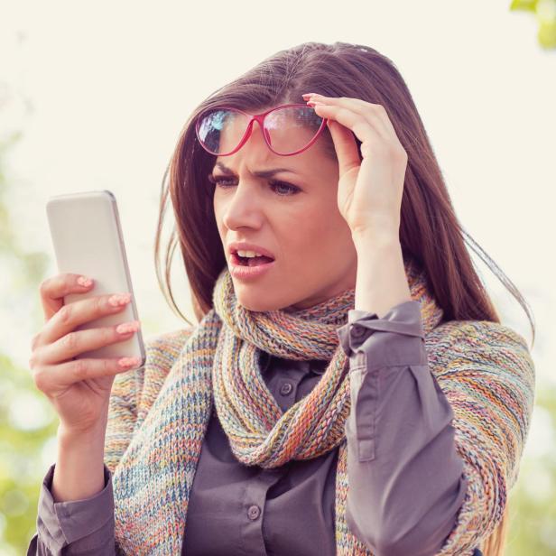 Annoyed upset woman in glasses looking at her smart phone with frustration while walking on a street on an autumn day