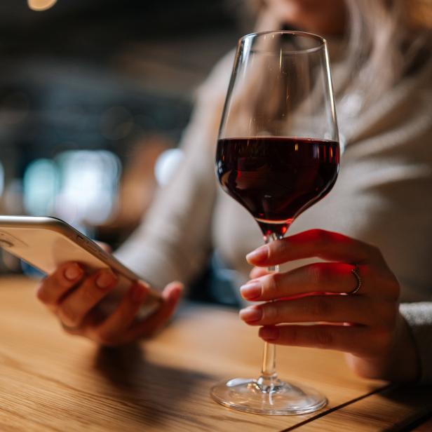 Eine Frau sitzt mit einem Glas Rotwein und einem Smartphone an einem Tisch.