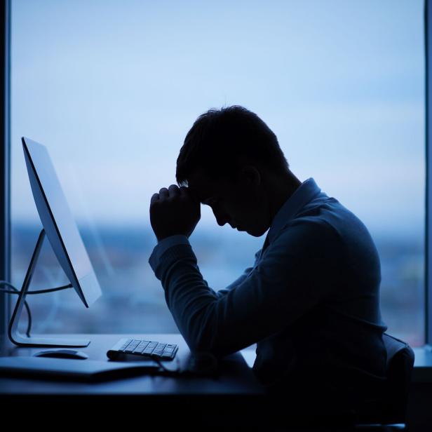 Eine Person sitzt mit gesenktem Kopf und verschränkten Händen nachdenklich vor einem Computer am Schreibtisch.