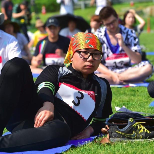 Eine Gruppe von Menschen sitzt auf Matten im Gras, einige tragen Startnummern.