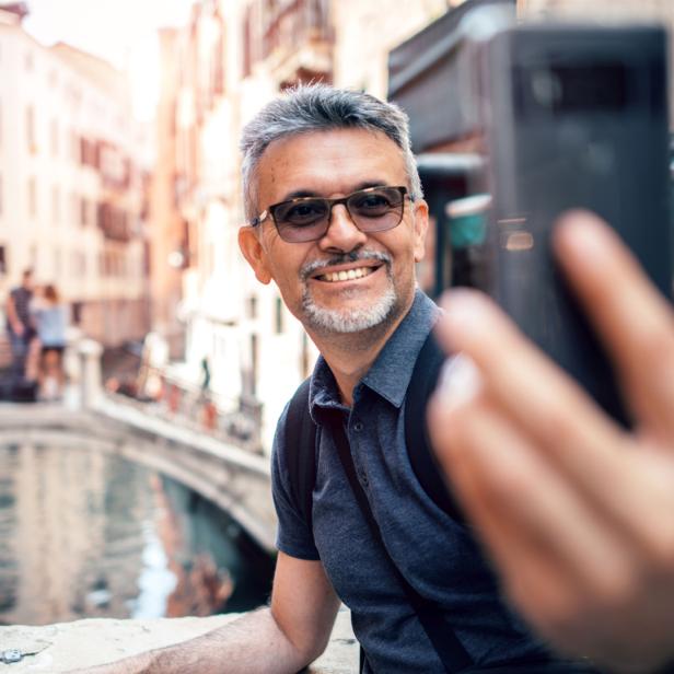 Tourist posiert für Selfie mit Smartphone vor einem Kanal mit Brücke darüber in Venedig