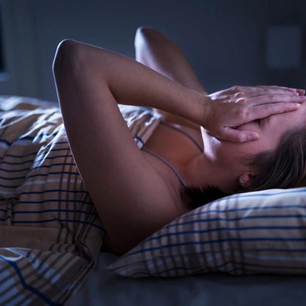 Eine Frau liegt im Bett und bedeckt ihr Gesicht mit den Händen, wirkt erschöpft oder gestresst.