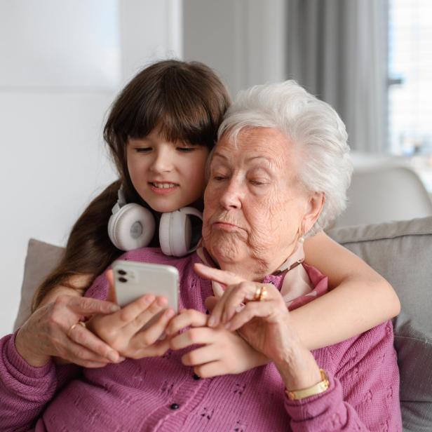 Grandmother with cute girl scrolling on smartphone, girl teaching senior woman to work with technology, internet. Portrait of elderly woman spending time with granddaughter.