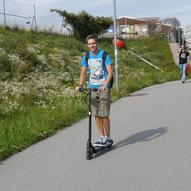 Ein junger Mann fährt mit einem Elektroroller auf einem Gehweg entlang.