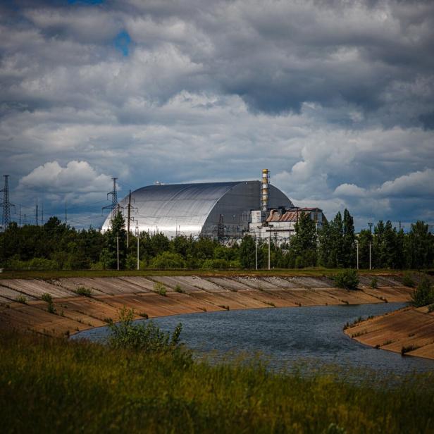 Der Sarkophag von Tschernobyl unter einem bewölkten Himmel, gesehen von einem grasbewachsenen Ufer aus.