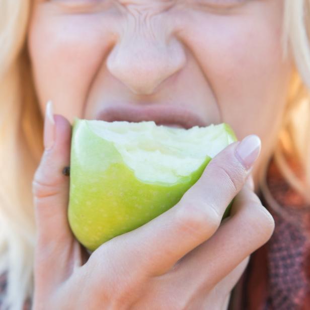 Eine blonde Frau beißt in einen grünen Apfel und verzieht dabei das Gesicht