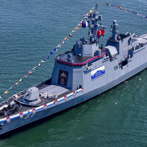 Ein großes graues Kriegsschiff mit nordkoreanischer Flagge fährt geschmückt mit bunten Wimpeln auf dem Wasser.