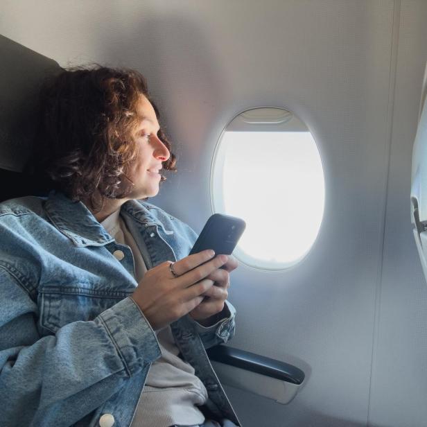 Passenger woman is flying in plane. Girl using phone sitting by airplane window. Traveling female with smartphone inside plane enjoying flight. Woman using wifi connection on board. Traveling girl.