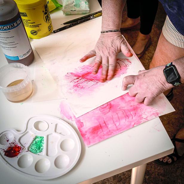 Eine ältere Frau malt mit den Fingern pinke Farbe auf Papier.