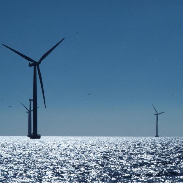 Windräder auf offener See, die Sonne spiegelt sich auf der Wasseroberfläche.