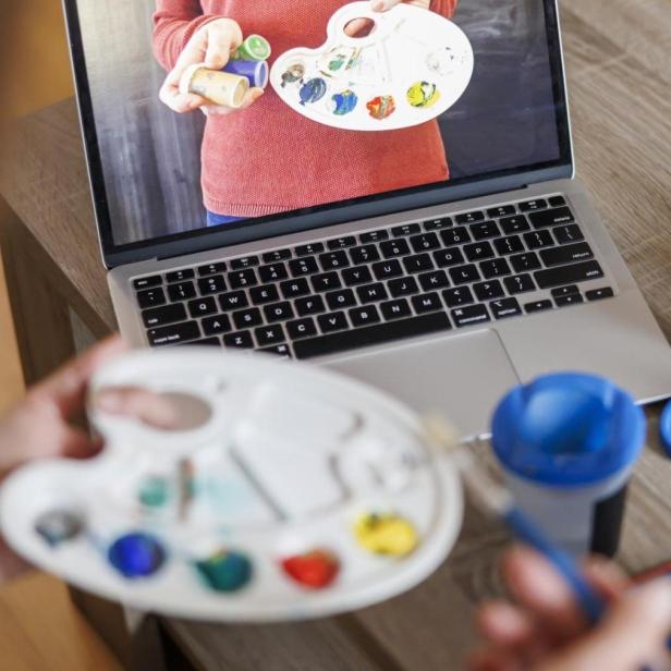 eine Person hält eine Malerpalette, im Hintergrund eine Leinwand und ein Laptop, auf dem ebenfalls eine Malerpalette zu sehen ist