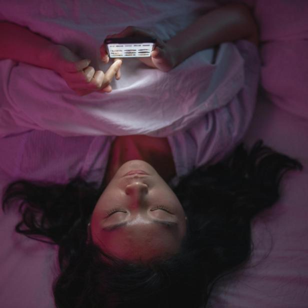 Eine Frau liegt auf ihrem Bett und scrollt auf ihrem Smartphone im Dunklen 