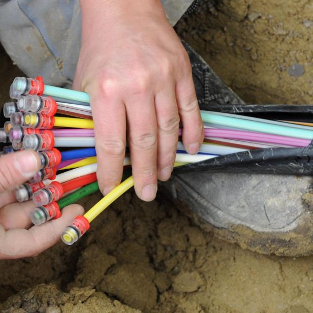 zwei Hände halten bunte Einzelstränge eines Glasfaserkabels in einer sandigen Grube