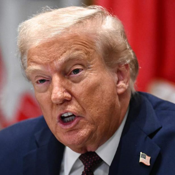 Donald Trump spricht ernsthaft, trägt einen dunklen Anzug mit US-Flaggen-Pin und sitzt vor roten Fahnen.