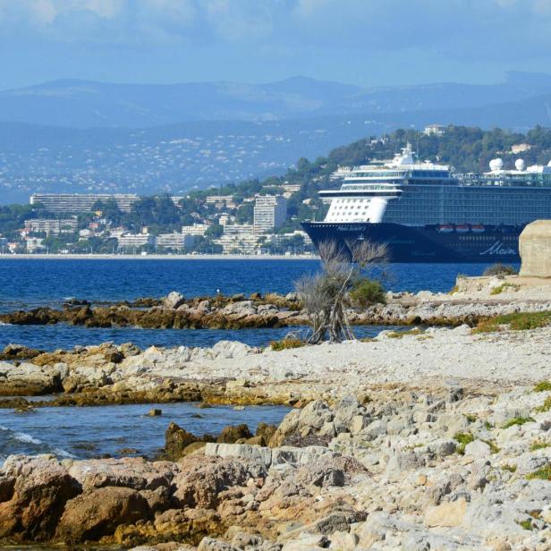 Eine felsige Küstenlinie mit schroffen Steinen und sanften Wellen, die gegen die Felsen schlagen, im Hintergrund ein Kreuzfahrtschiff.
