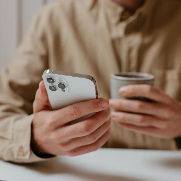 Ein Mann hält in der einen Hand ein iPhone und in der anderen einen Kaffee.