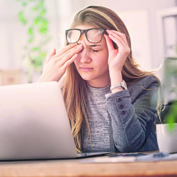 Eine junge Frau sitzt am Schreibtisch vor einem Laptop und reibt sich erschöpft die Augen.