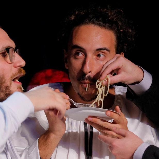 Zwei Männer essen gemeinsam Spaghetti von einem Teller, während mehrere Hände ihnen helfen.