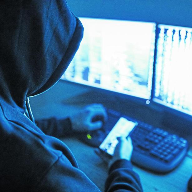 Eine Person mit Kapuze sitzt vor zwei hellen Monitoren und hält ein Smartphone in der Hand.