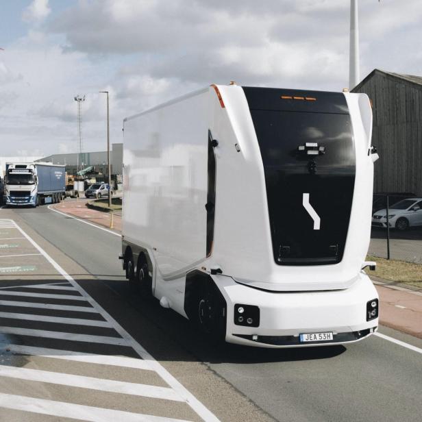 Ein weißer, autonomer Lkw mit schwarzer Front fährt auf einer Straße mit Windrädern im Hintergrund.