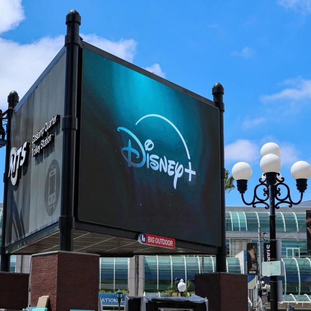 Zwei Personen arbeiten auf einer Hebebühne an einer großen Werbetafel mit Disney+ Logo an einer Straßenkreuzung.