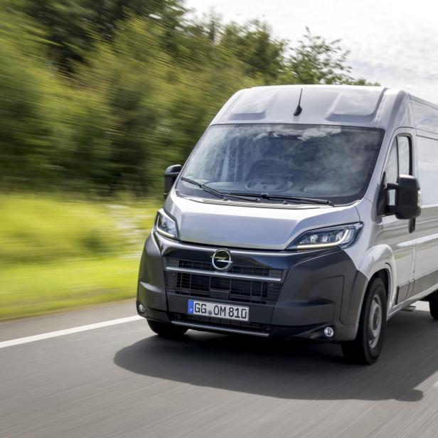 Ein Kastenwagen fährt auf einer Straße, im Hintergrund sieht man grüne Bäume.