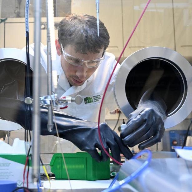 Ein Mann arbeitet mit Schutzhandschuhen in einer geschlossenen Laborapparatur an technischen Geräten.