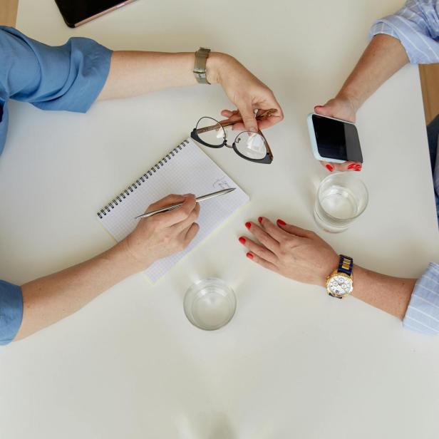 Zwei Personen sitzen an einem Tisch und unterhalten sich, eine Person hält ein Smartphone, die andere einen stift