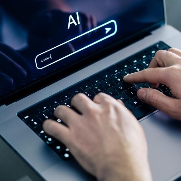Hände tippen auf einer Laptop-Tastatur, auf dem Bildschirm steht „AI“ mit einer Suchleiste darunter.
