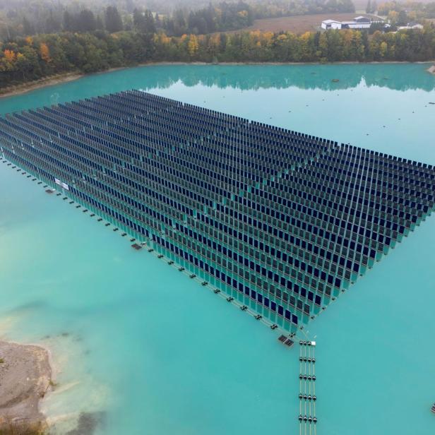 Eine große schwimmende Solaranlage befindet sich auf türkisfarbenem Wasser, umgeben von Bäumen und einer kleinen Landfläche.