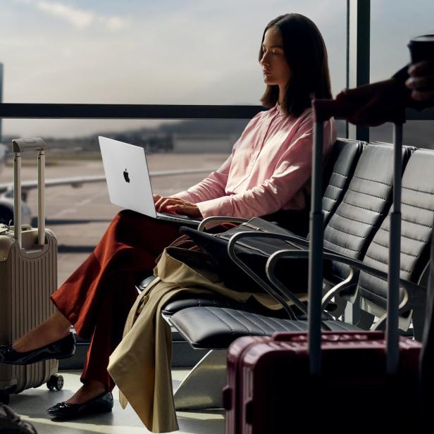 Frau arbeitet mit Laptop am Flughafen, Koffer neben ihr, Flugzeug im Hintergrund.