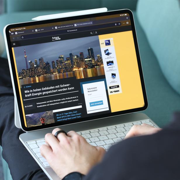 Eine Person sitzt auf einem Sofa und nutzt ein Tablet mit Tastatur, auf dem eine Technik-Webseite geöffnet ist.