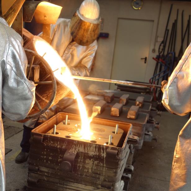 drei Menschen in silbernen Schutzanzügen gießen flüssiges Metall in eine Form