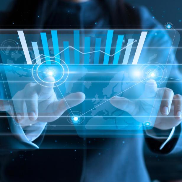 Person in Anzug tippt auf transparentes Display mit blauen Balkendiagrammen und Weltkarte im Hintergrund.