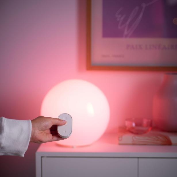 Hand hält Fernbedienung vor einer leuchtenden, kugelförmigen Lampe mit rosa Licht.