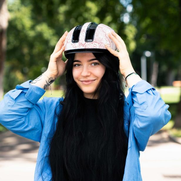 Eine junge Frau mit langen dunklen Haaren setzt sich im Park einen Fahrradhelm auf und lächelt.