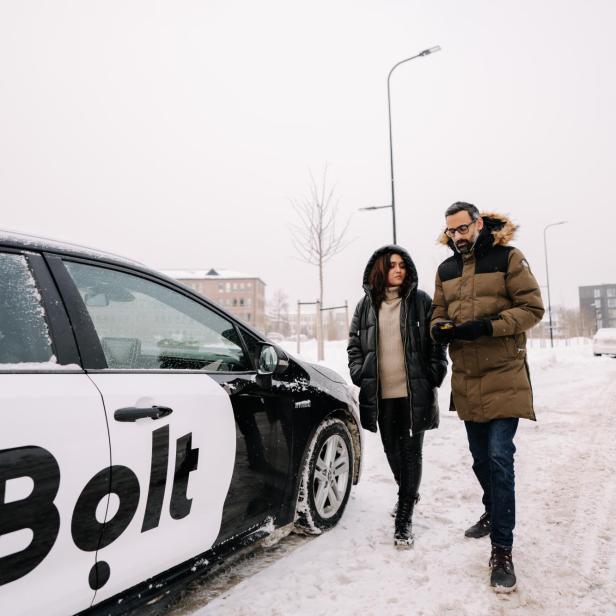 Zwei Personen gehen im Winter neben einem Bolt-Auto auf verschneiter Straße.