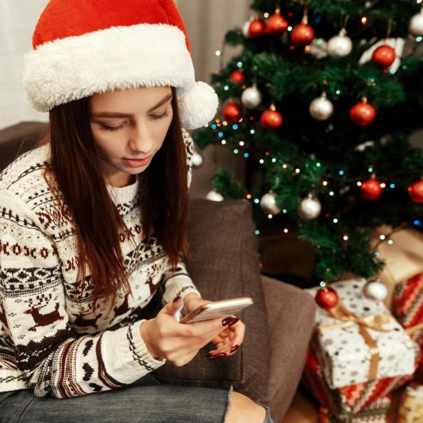 Frau mit weihnachtsmütze neben Christbaum und Geschenken schaut in ihr Smartphone