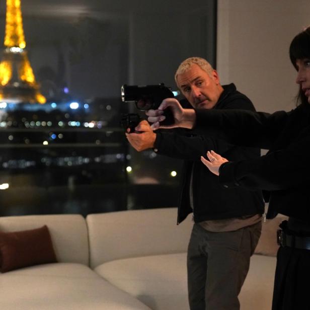 Zwei Ermittler aus dem Film „Zone 3“ zielen mit Waffen in einem modernen Wohnzimmer, im Hintergrund ist der beleuchtete Eiffelturm zu sehen.