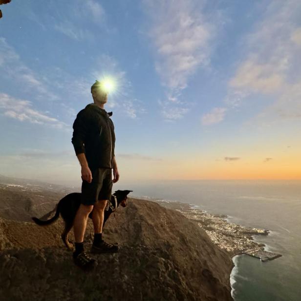 Mann mit Stirnlampe und Hund steht auf einer Klippe in der Dämmerung