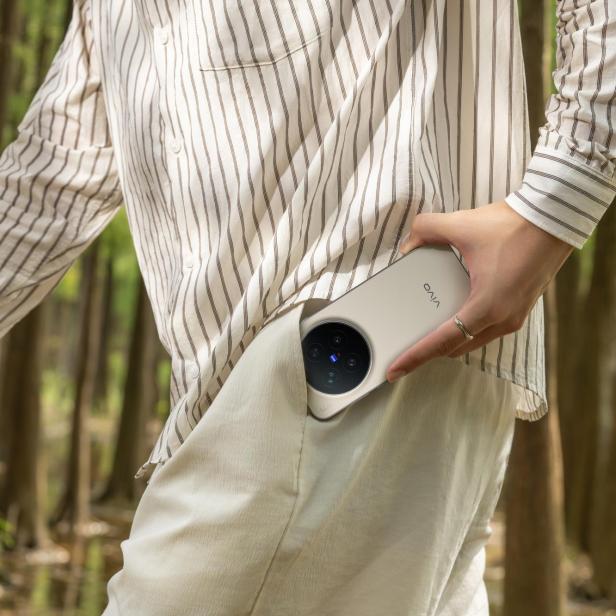 Eine Person steckt ein Smartphone mit großem Kameramodul in eine Hosentasche, während sie durch einen Wald geht.